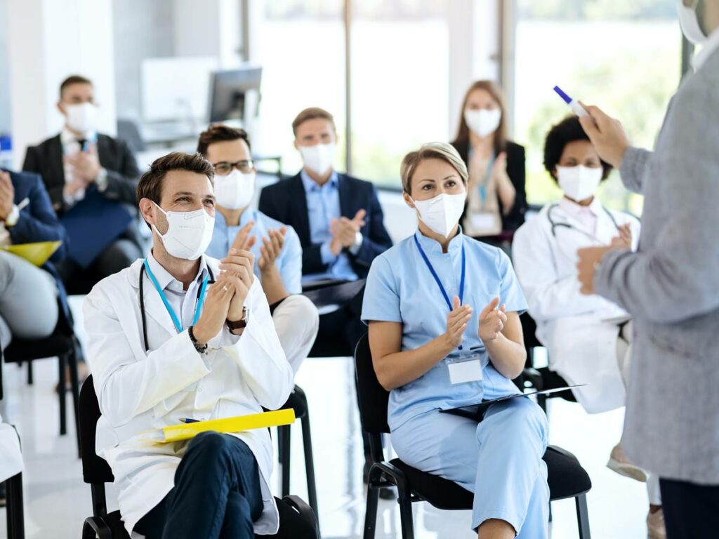 Large group of doctors and business people applauding while attending educational event during coronavirus pandemic. Focus is on male doctor.