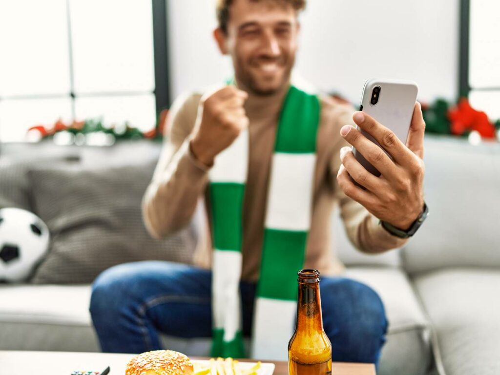 Young hispanic man supporting soccer match using smartphone sitting by christmas decor at home