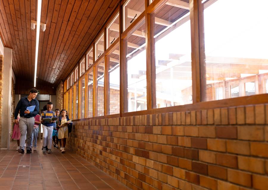 Diverse male teacher and children walking together in corridor at elementary school, copy space. School, learning, childhood, teaching and education, unaltered.