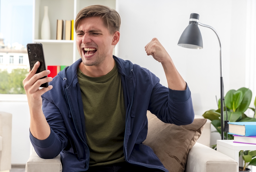 excited young blonde handsome man sits on armchair keeping fist and looking at phone inside design living room