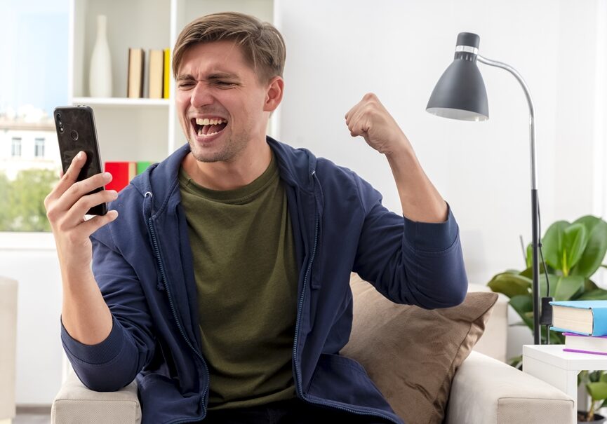 excited young blonde handsome man sits on armchair keeping fist and looking at phone inside design living room